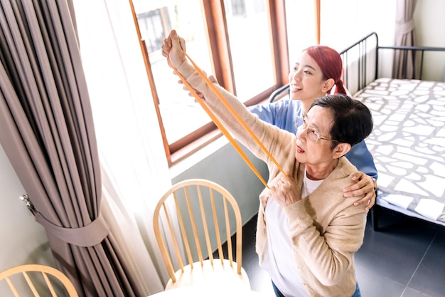 Pflegekraft, die Scrubs trägt, trainiert mit einer älteren asiatischen Frau, indem sie Widerstandsbandübungen für den älteren Patienten in der Physiotherapie-Behandlung verwendet Häusliches Gesundheits- und Pflegeheimkonzept