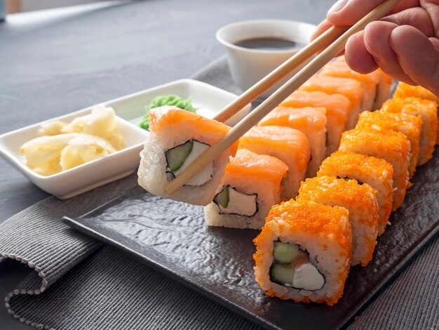 Philadelphia- und California-Rollen auf einem strukturierten dunklen Teller. Eine Rolle wird in Granny-Sticks über den anderen gehalten. Neben Sojasauce, Wasabi und Ingwer. Seitenansicht. japanische Küche