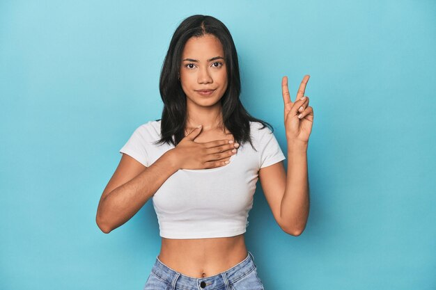 Philippinische junge Frau im blauen Studio, die einen Eid ablegt und die Hand auf die Brust legt