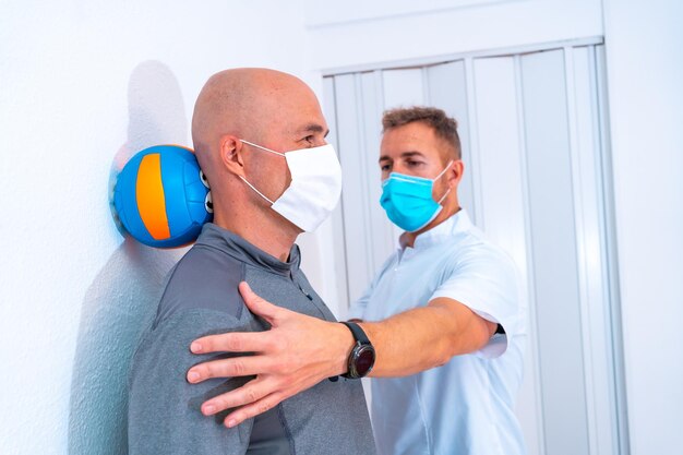 Physiotherapeut mit Maske hilft dem Kunden, den Nacken mit einem Haar an der Wand zu dehnen Physiotherapie mit Schutzmaßnahmen bei der Coronavirus-Pandemie COVID19 Osteopathie-Sport-Quiromassage