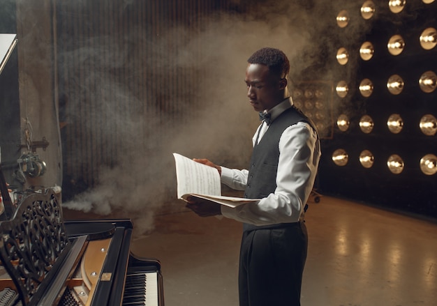 Pianista de ébano con cuaderno de música en sus manos en el escenario con focos. Artista posa en instrumento musical antes del concierto