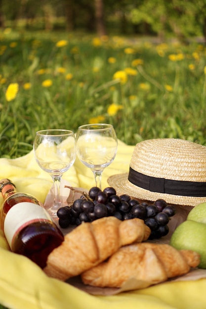 Picknick auf dem Gras mit Croissant, rosa Wein, Strohhut, Traube auf gelbem Plaid und grünem Gras. Sommerzeit - vertikales Foto