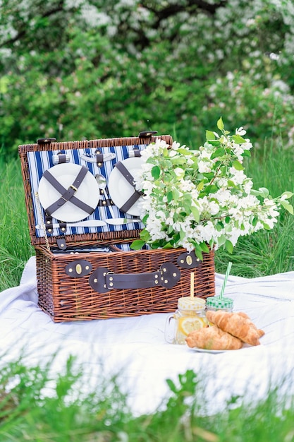 Picknickkorb mit Limonade und Croissants auf einer weißen Decke in einem blühenden Apfelgarten Vertikales Foto