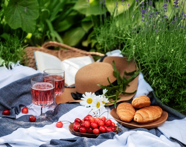 Picnic al aire libre en campos de lavanda. Vino rosado en una copa, cerezas y sombrero de paja sobre una manta