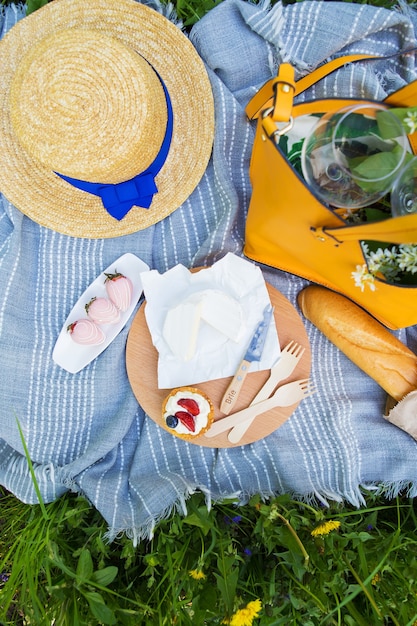 Picnic brillante en la naturaleza, queso brie en el tablero y pastel con fresas, sombrero, bolsa en la hierba