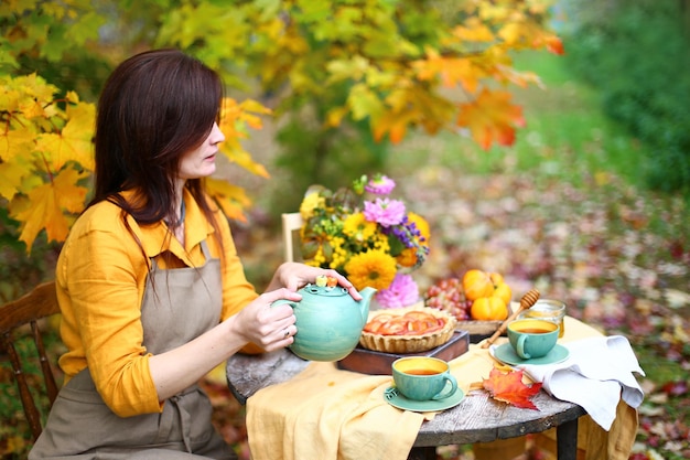 Picnic de otoño Mujer con vestido amarillo y delantal de lino bebe té de una taza en una mesa de madera en el jardín Hermoso mantel de tetera miel con cuchara pastel de manzana cosecha caqui uvas hoja de arce