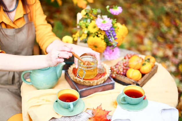 Picnic de otoño Mujer con vestido amarillo y delantal de lino bebe té de una taza en una mesa de madera en el jardín Hermoso mantel de tetera miel con cuchara pastel de manzana cosecha caqui uvas hoja de arce