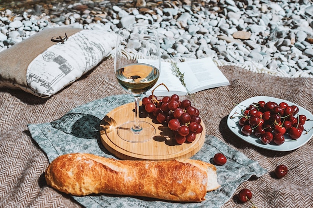 picnic de verano en la costa con copa de vino blanco baguette cerezas uva libro abierto almohada