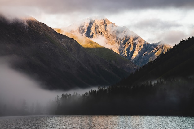 Pico de alta montaña de oro y muchas nubes bajas sobre el lago de la montaña al amanecer. Niebla densa sobre el agua y bosque en hora dorada. Paisaje de montaña atmosférica temprano en la mañana. Paisaje relajante alpino.