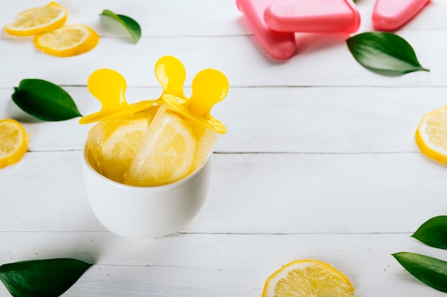 Picolés de limão em um copo sobre uma mesa de madeira branca decorada com rodelas de limão e folhas da natureza