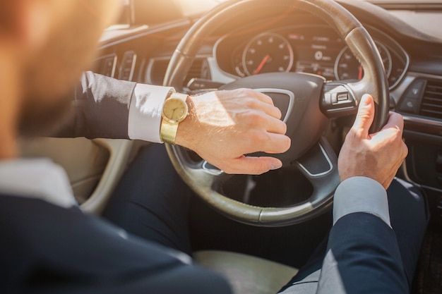 Picutr ofman sentado en el coche y mira los relojes. Él sostiene una mano en el volante. Está soleado afuera.