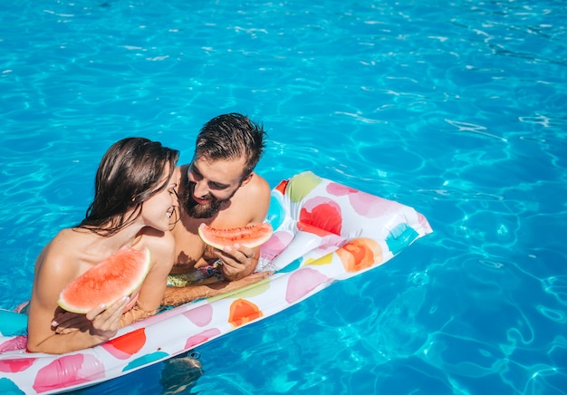 Foto picutre bonito de jovem e mulher, inclinando-se para colchão de ar e segurar pedaços de melancia. eles olham para o outro e sorriem. casal está na piscina.