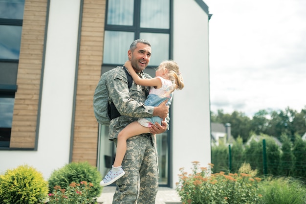 De pie cerca de la casa. Militar en uniforme se siente increíble de pie cerca de la casa con la hija