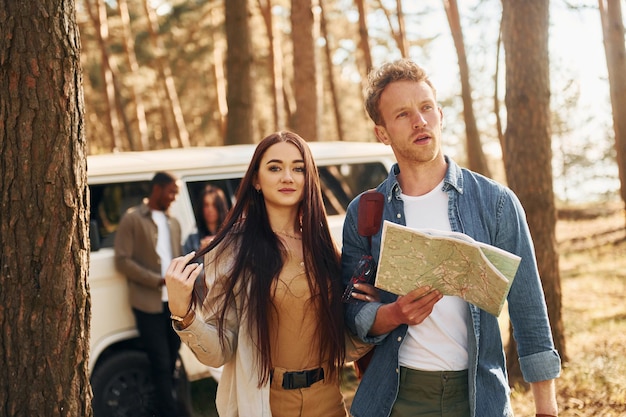 De pie cerca del coche Grupo de jóvenes viaja juntos en el bosque durante el día