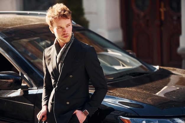 De pie cerca de coche negro Elegante joven en ropa elegante formal al aire libre en la ciudad