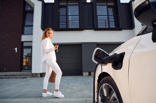 De pie cerca del edificio Mujer joven vestida de blanco está con su coche eléctrico durante el día