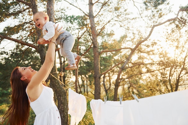 De pie cerca de la ropa blanca que cuelga de la cuerda para secarse Joven madre con su pequeño hijo está al aire libre en el bosque Hermoso sol