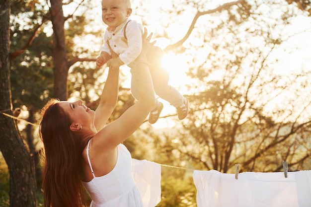 De pie cerca de la ropa blanca que cuelga de la cuerda para secarse Joven madre con su pequeño hijo está al aire libre en el bosque Hermoso sol