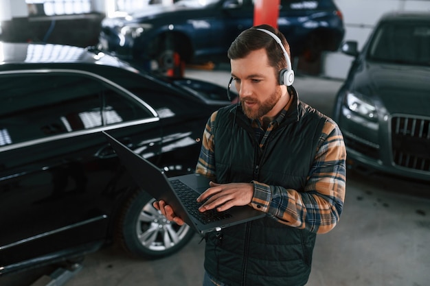 De pie con una computadora portátil en las manos y auriculares Un hombre en uniforme está trabajando en el salón de automóviles