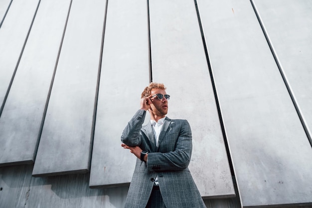 Foto de pie contra la pared del edificio un joven hombre de negocios con ropa formal gris está al aire libre en la ciudad