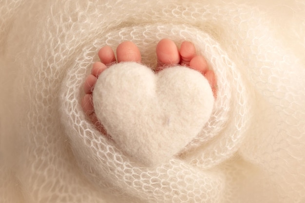 El pie diminuto de un bebé recién nacido. Pies suaves de un recién nacido en una manta de lana blanca. Cerca de los dedos de los pies, los talones y los pies de un recién nacido. Corazón blanco tejido en las piernas de un bebé. Fotografía macro de estudio.