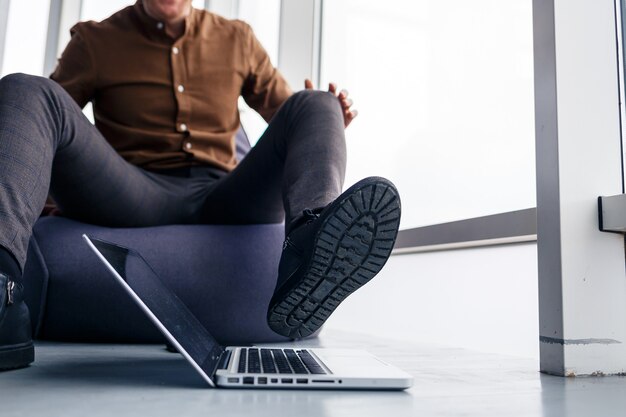 El pie de un hombre con zapatos está cerca de la computadora portátil. El hombre quiere pisar su computadora portátil