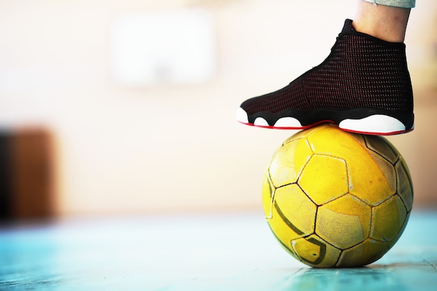 Un pie humano descansa sobre el balón de fútbol en el piso de concreto. Foto de una pelota de fútbol y zapatillas en un piso de madera.