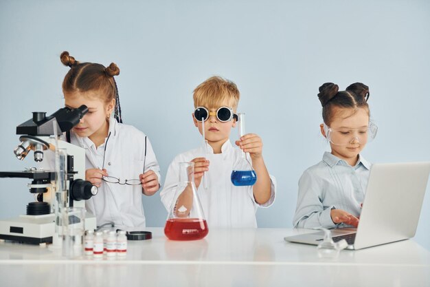 De pie junto a la mesa con tubos de ensayo Los niños con batas blancas juegan a los científicos en el laboratorio usando equipos