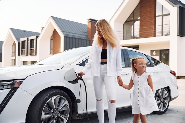 De pie juntos Mujer joven con su pequeña hija está con su coche eléctrico al aire libre