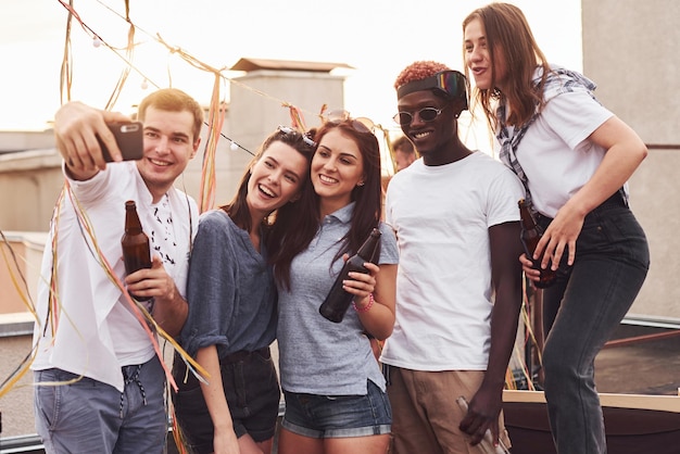 De pie juntos y tomando selfie Grupo de jóvenes con ropa informal tienen una fiesta en la azotea juntos durante el día