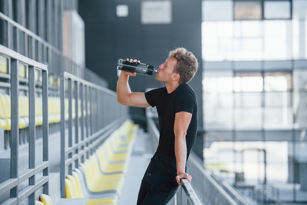 De pie y sosteniendo una botella de agua Un joven deportivo con camisa negra y pantalones al aire libre durante el día