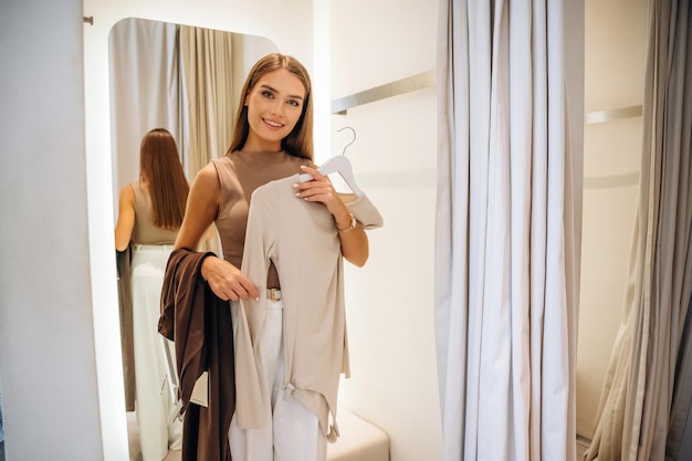 De pie sosteniendo suéter y pantalones sonriendo Una mujer se prueba ropa en una tienda en un camerino