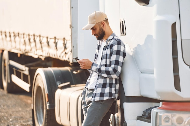 De pie y sosteniendo el teléfono inteligente El joven camionero está con su vehículo durante el día