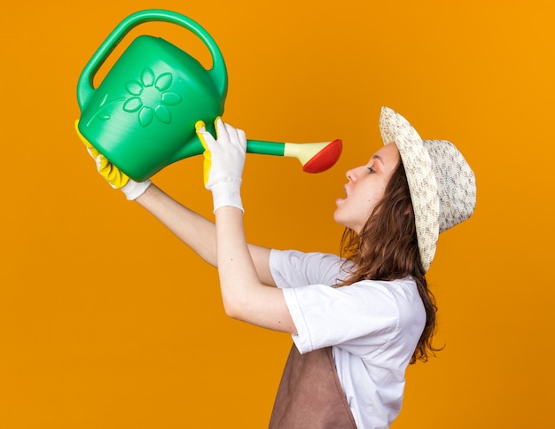 De pie en la vista de perfil joven jardinera con sombrero y guantes de jardinería que se riega con regadera aislada en la pared naranja