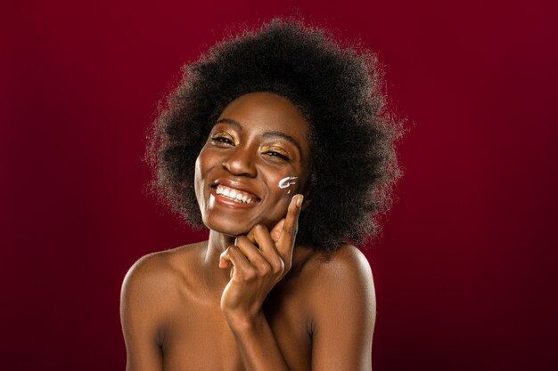 Para la piel. Encantado de mujer afroamericana sonriendo mientras se pone crema en la mejilla