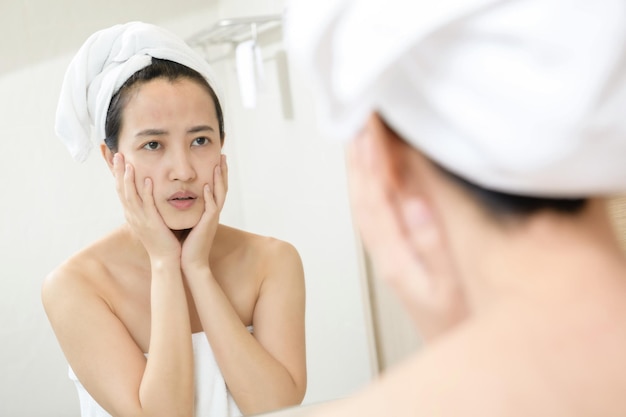 Piel problemática Mujeres asiáticas jóvenes preocupadas haciendo estallar un grano en la mejilla mientras están de pie cerca del espejo en el baño mujeres asiáticas jóvenes con acné