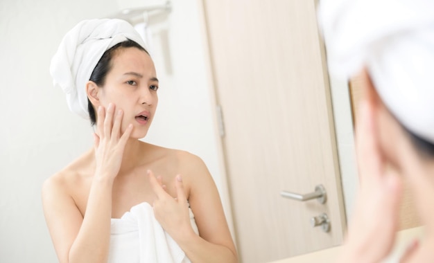 Piel problemática Mujeres asiáticas jóvenes preocupadas haciendo estallar un grano en la mejilla mientras están de pie cerca del espejo en el baño mujeres asiáticas jóvenes con acné