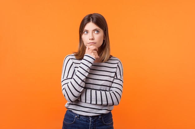 Piensa en un plan. Retrato de mujer pensativa con cabello castaño en camisa a rayas de manga larga de pie sosteniendo la mano en la barbilla y mirando hacia arriba, pensando intensamente. tiro de estudio interior aislado sobre fondo naranja