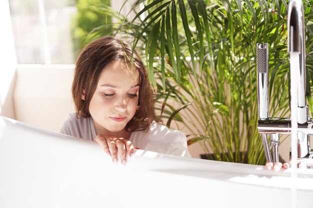 Piensa positivo. Chica en un baño luminoso con una planta verde. Una mujer feliz espera hasta que la bañera se llene de agua.