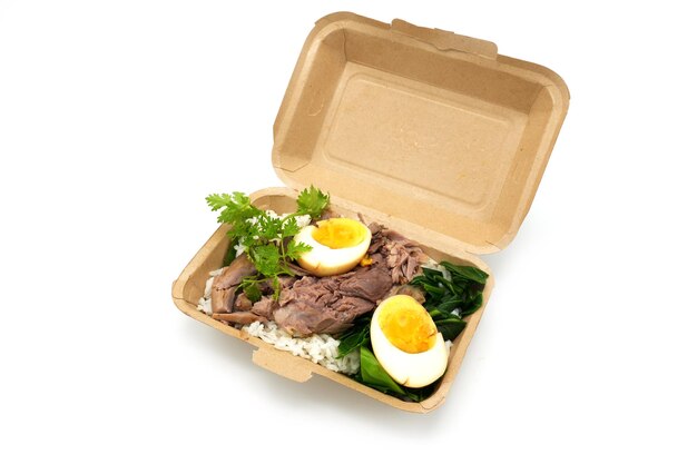 Pierna de cerdo guisada con huevo cocido y verduras sobre arroz en una caja desechable marrón sobre fondo blanco.