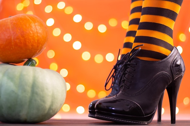 Piernas de bruja en medias a rayas y zapatos de tacón con calabazas de Halloween sobre un fondo naranja con bokeh. Copie el espacio.