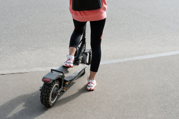 Las piernas de una chica desconocida con zapatillas blancas y mallas de gimnasia montada en un scooter eléctrico negro sobre asfalto urbano. Transporte moderno, patinete eléctrico.