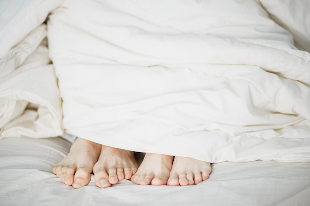 Las piernas de dos amantes, hombres y mujeres, niñas y niños, sobresalen debajo de la manta blanca, suave, esponjosa y limpia en la cama, el primer plano blanco