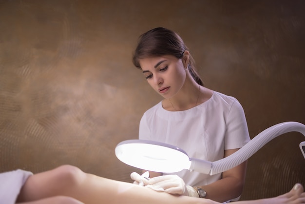 Piernas femeninas en hoja morada durante la depilación por una esteticista profesional con guantes. Spa, industria de la belleza, tratamiento en clínica, electrólisis.