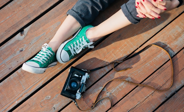 Piernas femeninas en zapatillas hipster y cámara retro en la superficie de madera al aire libre, fotos de cultivos