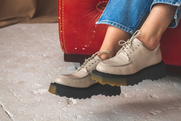 Piernas femeninas con zapatos de cuero. Primer plano de zapatillas adolescentes. Chica adolescente sentada en una silla de cuero vieja en el estudio, fondo de hormigón