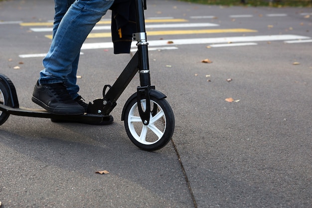 Las piernas de un hombre en jeans y zapatillas de deporte en un scooter en el parque en otoño con hojas amarillas secas caídas sobre el asfalto. Paseos de otoño, estilo de vida activo, transporte ecológico, tráfico