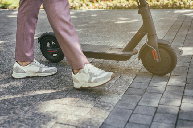 Piernas del hombre que van cerca de scooter eléctrico ecológico en baldosas de acera de la ciudad en un día soleado