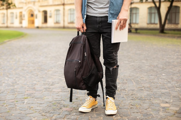 Piernas de hombre en zapatillas amarillas, con camisa gris, jeans ajustados negros en el campus universitario en otoño