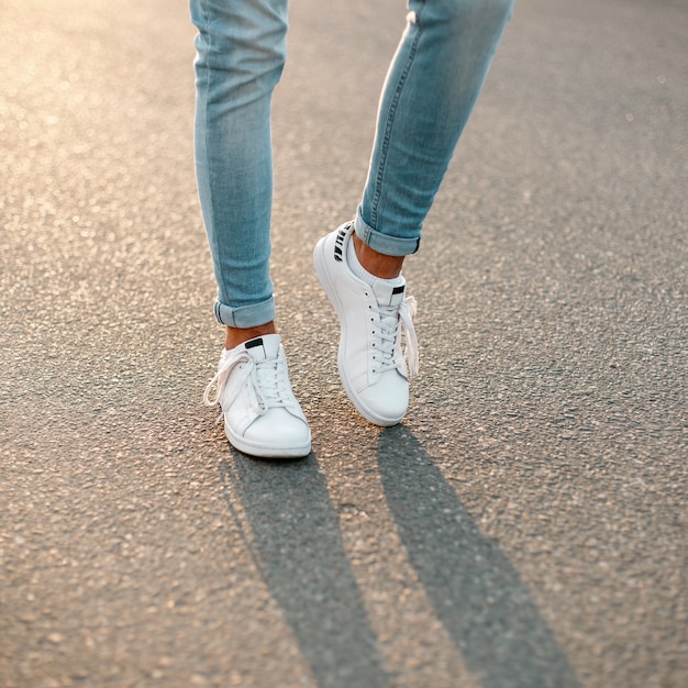 Hombre joven moderno de moda en jeans con estilo en unas zapatillas blancas  de temporada de cuero de moda se encuentra en un estacionamiento en la  ciudad. primer plano de las piernas